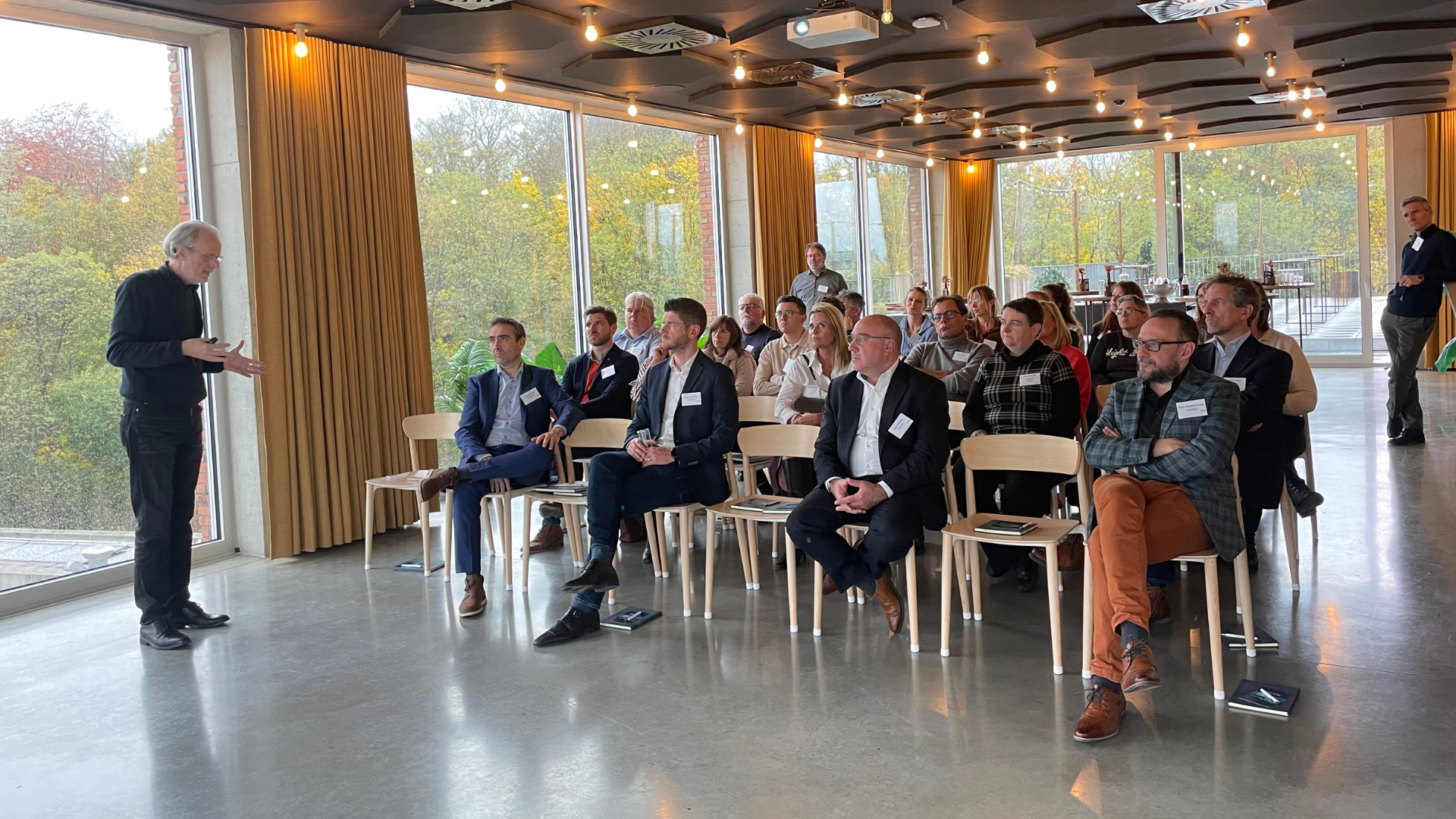 Een spreker presenteert voor een groep aandachtige deelnemers tijdens een zakelijk event, met grote ramen en uitzicht op een groene omgeving.