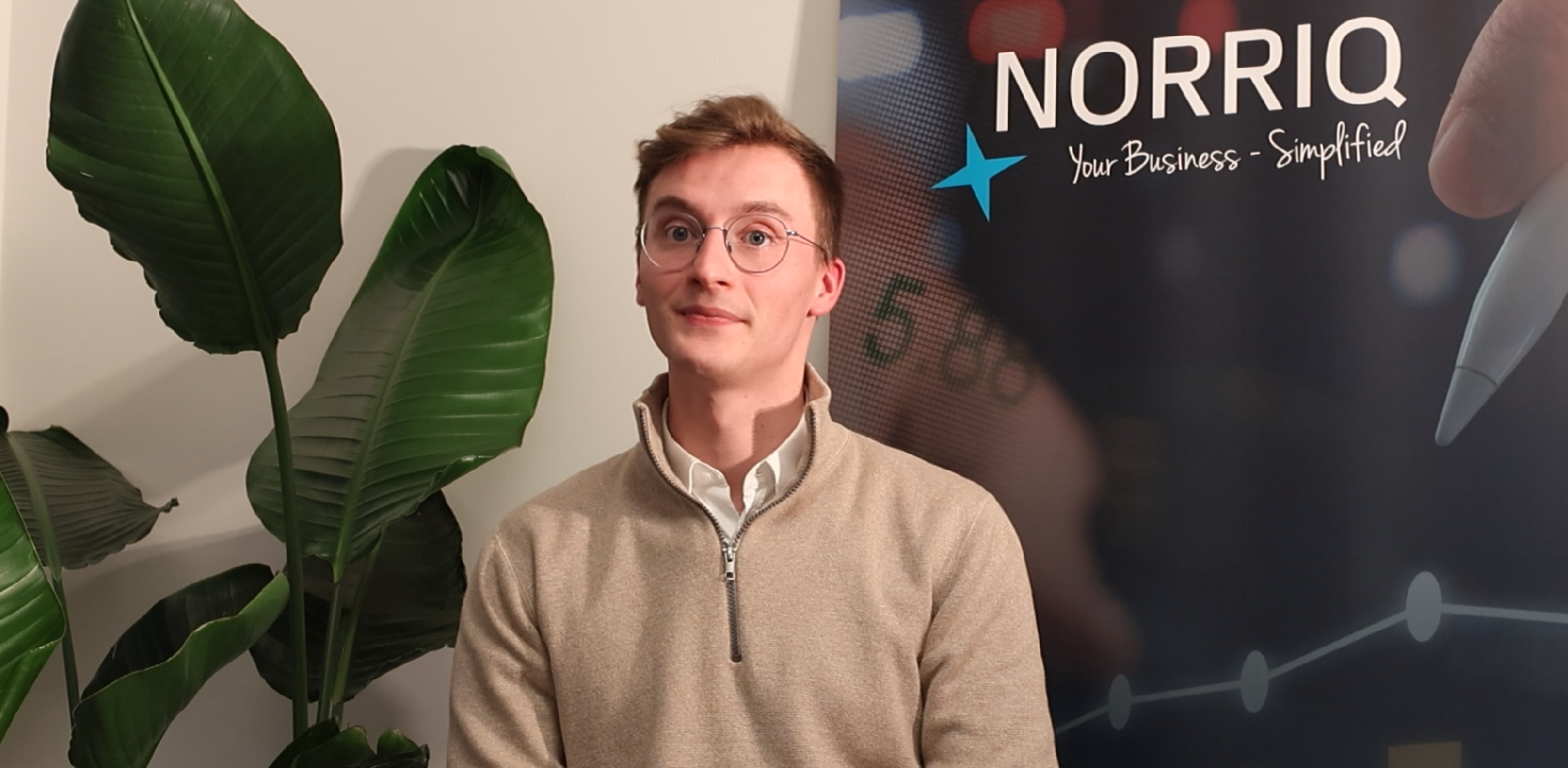 A man sitting near a plant and a roll-up which is branded NORRIQ