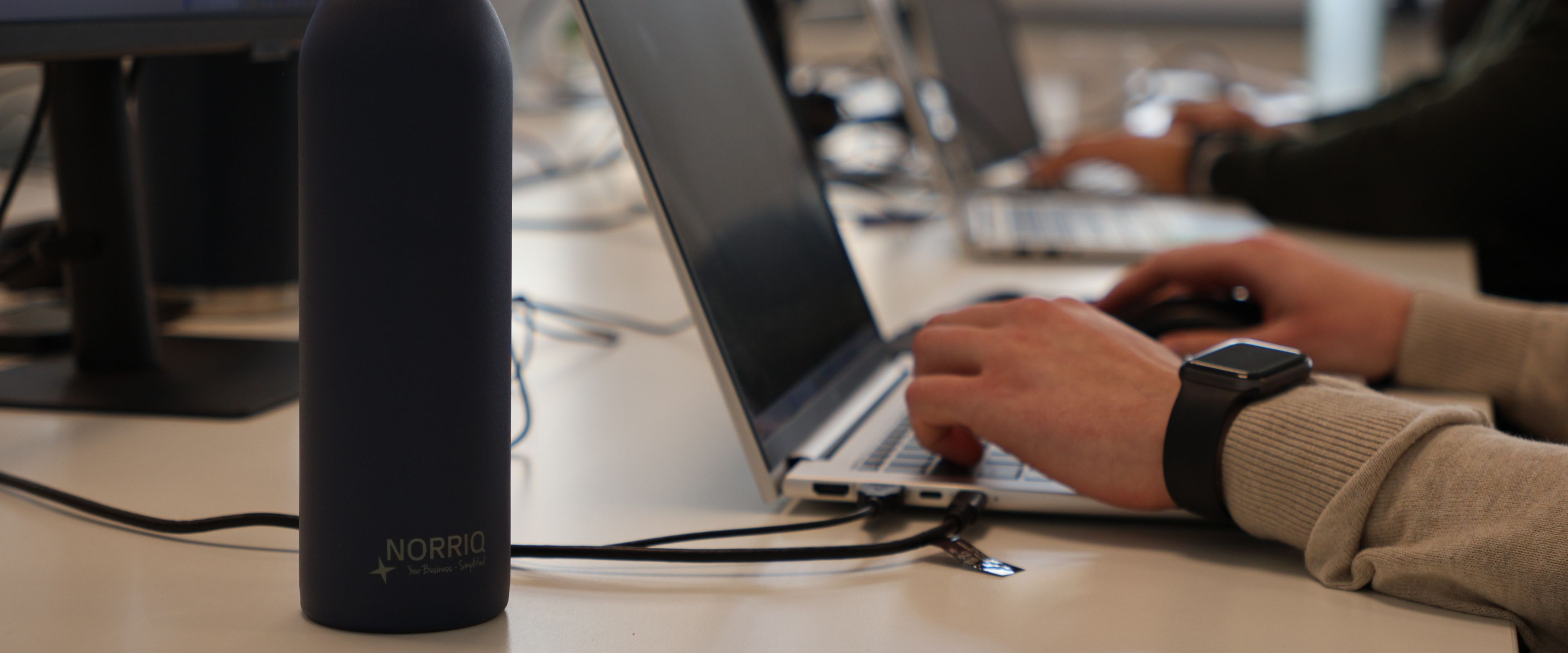 Hands typing on a laptop in an office with a NORRIQ water bottle on the desk.
