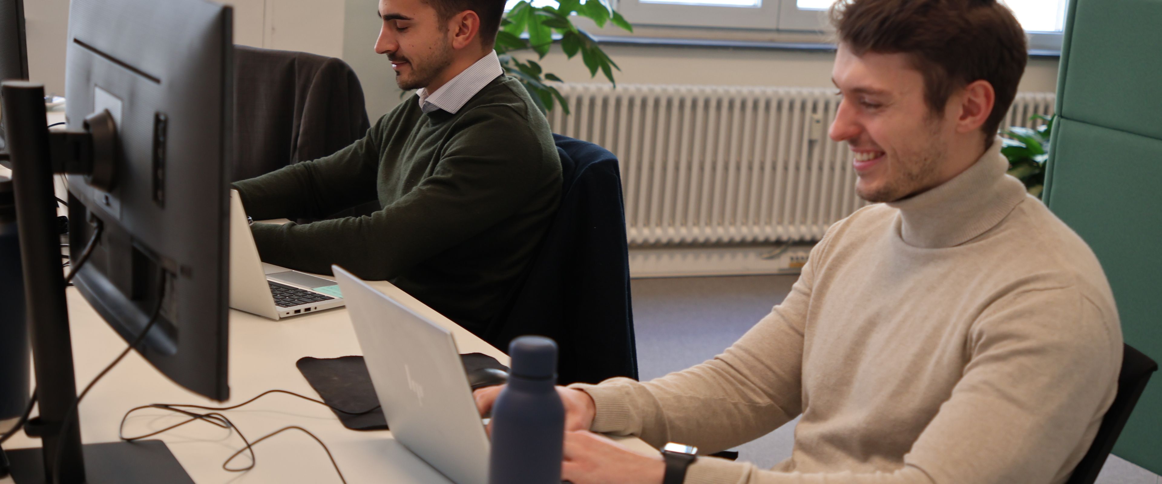 Two people working on laptops at a desk with a water bottle nearby.